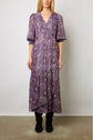 Woman wearing a long, flowy purple robe fleurie with a patterned design, three-quarter puff sleeves, and black boots, standing against a plain white background.