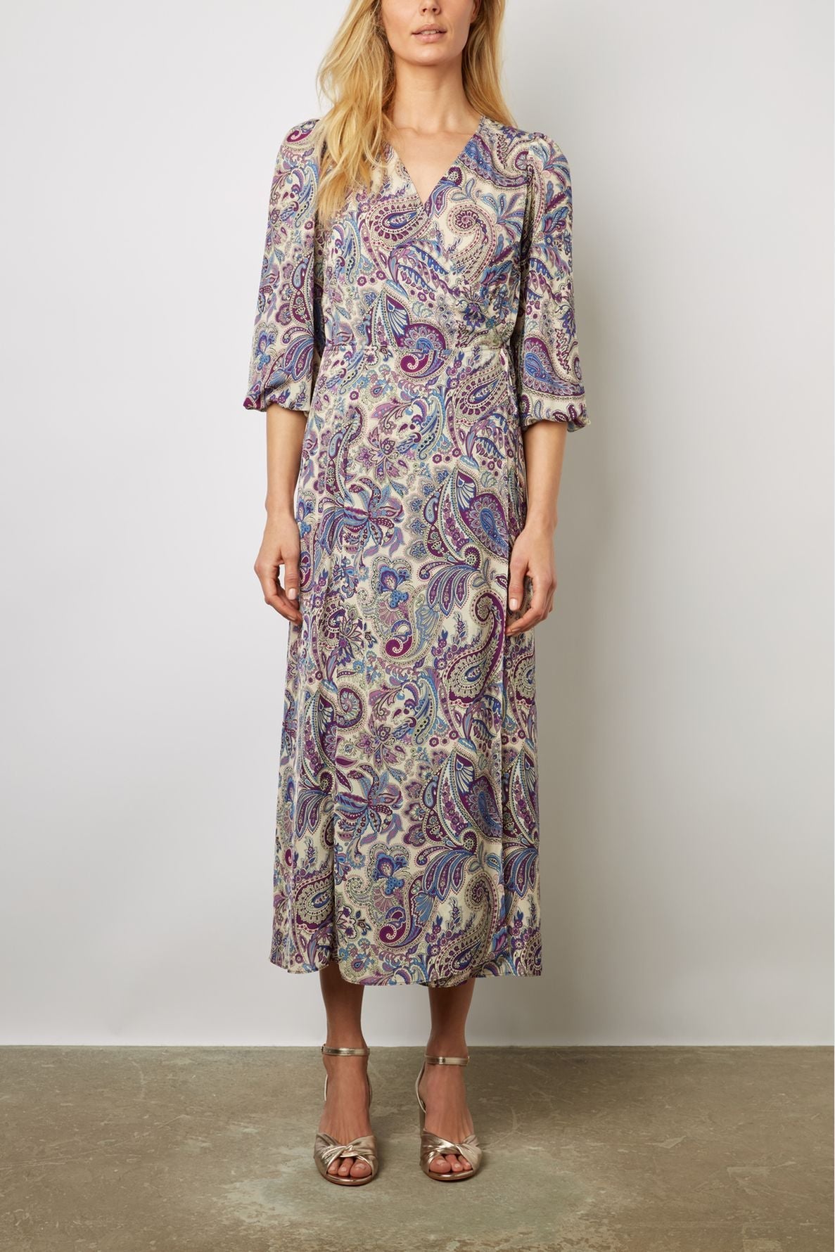 Woman wearing a long-sleeve, V-neck, ankle-length robe portefeuille with a purple and blue imprimé cachemire, standing against a plain light background, paired with silver open-toe heels.