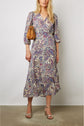 Woman wearing a long-sleeved, imprimé cachemire robe portefeuille with a brown handbag and metallic strappy sandals, standing against a plain wall.