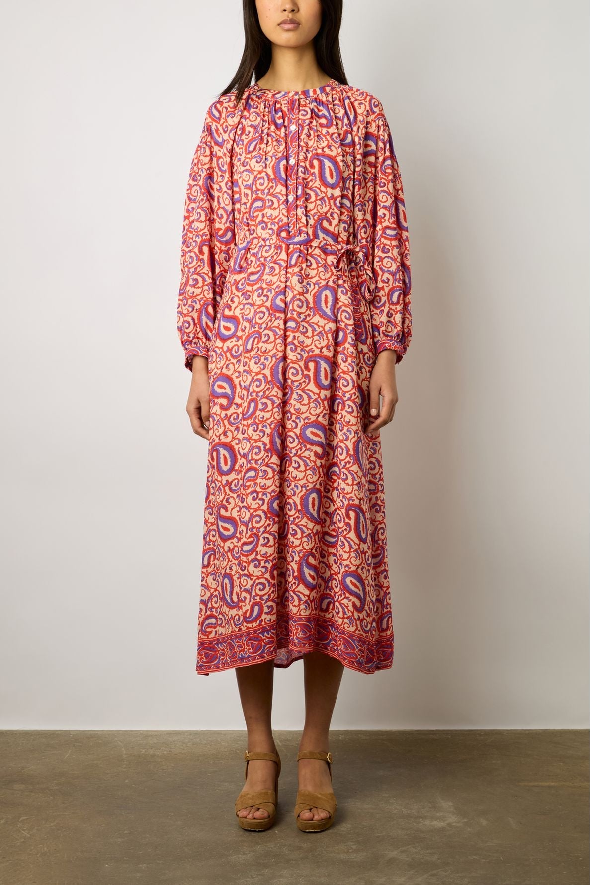 A woman wearing a long-sleeved, ankle-length robe longue fluide with a red and pink imprimé ethnique stands against a plain white wall, wearing tan heeled sandals.