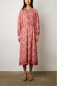 A woman wearing a long-sleeved, ankle-length robe longue fluide with a red and pink imprimé ethnique stands against a plain white wall, wearing tan heeled sandals.