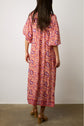 A woman stands facing away, wearing a robe longue fluide with an imprimé ethnique in pink and orange paisley, featuring elbow-length sleeves and brown heeled sandals, from the collection Studio, set against a plain background.