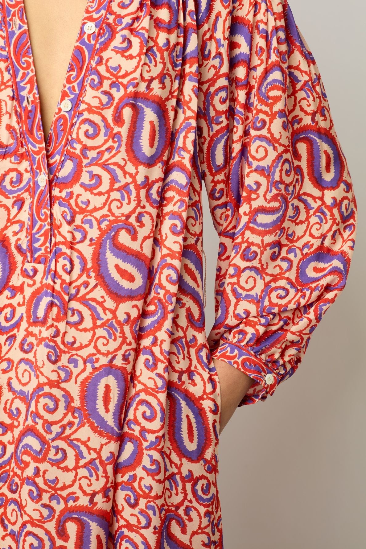 Person wearing a colorful, paisley-patterned garment in red, purple, and white, with a V-neckline and loose sleeves. This robe longue fluide from the collection Studio features an élégant imprimé ethnique. Only the left side of the upper body and part of the arm are visible.