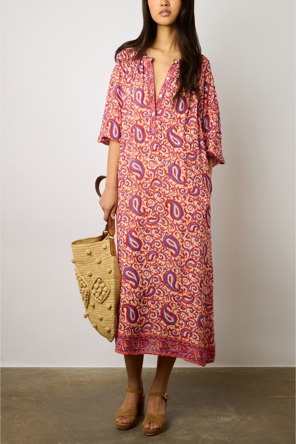 A woman wearing a pink-and-red patterned robe longue fluide with un imprimé ethnique from the collection Studio holds a woven straw bag and wears tan platform sandals, standing against a plain background.