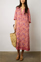 A woman wearing a pink-and-red patterned robe longue fluide with un imprimé ethnique from the collection Studio holds a woven straw bag and wears tan platform sandals, standing against a plain background.
