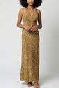 A person wearing a sleeveless, robe longue halter-neck, floor-length dress with a leopard print pattern and tan sandals stands against a plain light gray background.