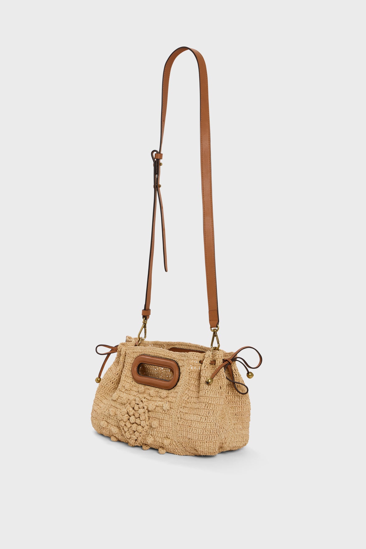 A beige woven straw sac à main, crafted from raphia naturel in an artisanal style, features a long adjustable brown leather strap, wooden oval handle, and drawstring sides. It is displayed against a plain white background.