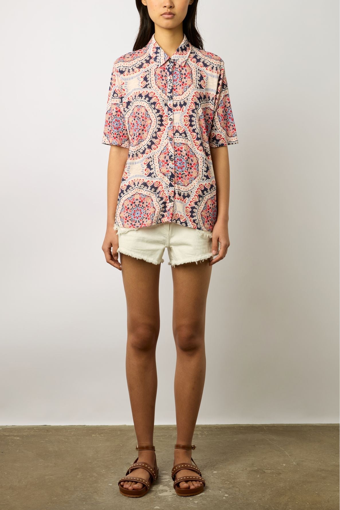 A person wearing a pink patterned short-sleeve button-up shirt, white frayed shorts, and brown sandals stands against a plain light-colored wall.