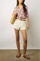 Woman wearing a colorful patterned shirt, white high-waisted shorts, brown sandals, and carrying a woven beige shoulder bag, standing against a plain background.