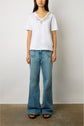 A woman stands against a plain background, wearing a white T-shirt col V en coton adorned with floral embellishments and delicate strass accents, light blue wide-leg jeans, and black sandals. Her arms rest by her sides.