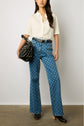 A woman stands against a plain wall, wearing a white short-sleeve shirt, blue patterned high-waisted jeans, black heeled clogs, and holding a black handbag.