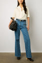 A woman stands against a plain wall, wearing a white short-sleeve shirt, high-waisted blue patterned jeans, black clogs, and holding a black handbag.