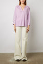 A person wearing a loose lavender T-shirt lin with col tunisien and wide-leg off-white pants stands against a plain white background, facing forward with arms relaxed at their sides.