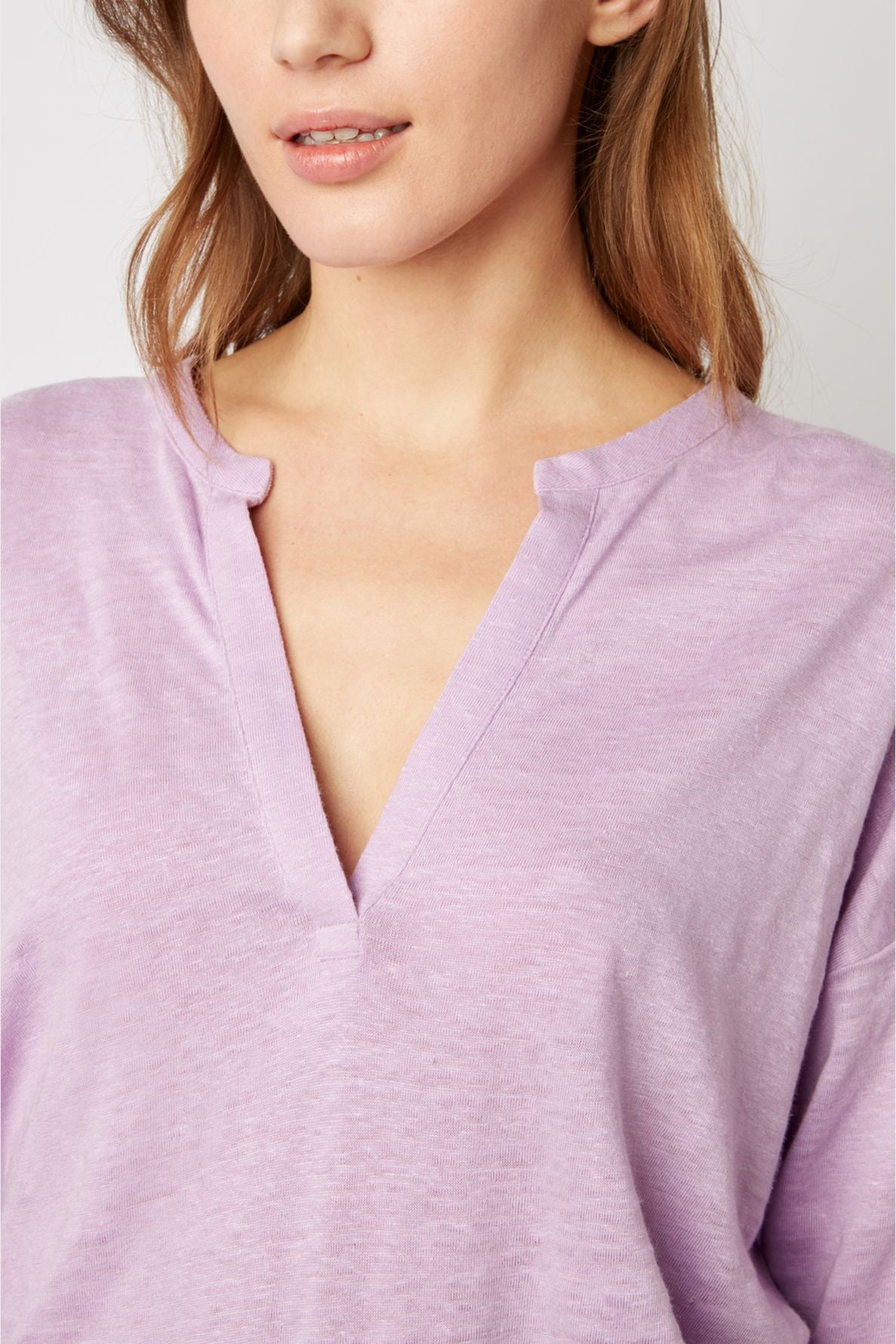 A woman with light brown hair wears a light purple V-neck top featuring a unique notch at the neckline reminiscent of a col tunisien. The image is cropped to show her from the lips to the chest.