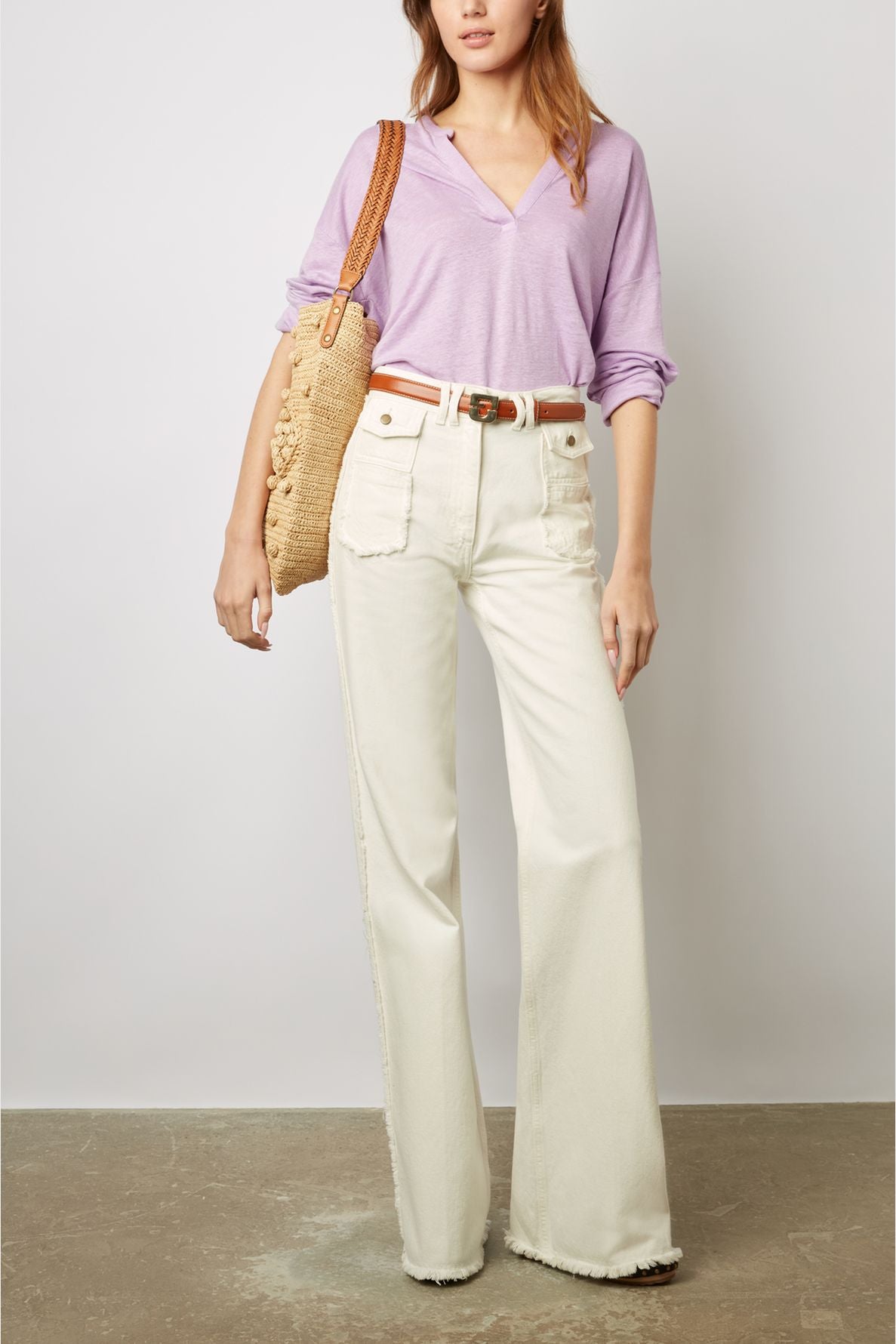 Woman wearing a light purple T-shirt en lin à col tunisien et manches 3/4, cream wide-leg jeans with front pockets, a brown belt, and carrying a woven straw shoulder bag, standing against a plain background.
