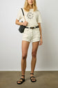 Woman wearing a white graphic T-shirt, white distressed denim shorts, black belt, black shoulder bag, and black studded sandals, standing against a plain wall.
