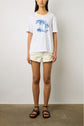 A person stands against a plain wall wearing a white T-shirt with blue palm tree print, light frayed shorts, and black sandals.
