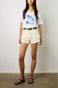 A woman wearing a white graphic t-shirt with palm trees, cream high-waisted shorts with large pockets, a black belt, and black sandals standing against a plain background.