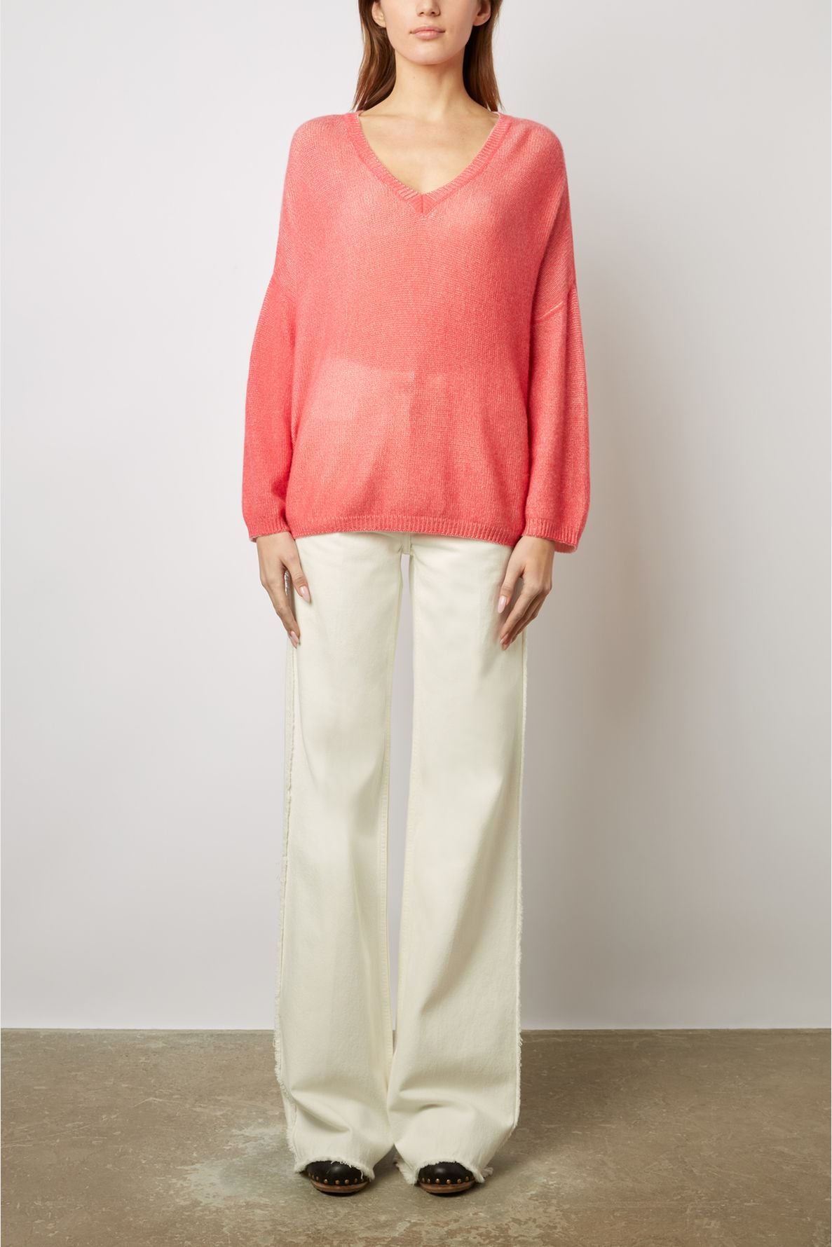 A person stands against a plain background wearing a loose-fitting pink V-neck sweater and wide-leg white pants, with their hands by their sides. Their face is partially visible and their shoes are dark with a studded design.