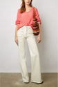 A person wearing a coral V-neck sweater, high-waisted white wide-leg jeans, and carrying a brown and red striped crochet shoulder bag stands against a plain background.