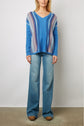 Woman wearing a blue V-neck pull cachemire with rétro multicolored rayé stripes and wide-leg blue jeans, standing against a plain light background.