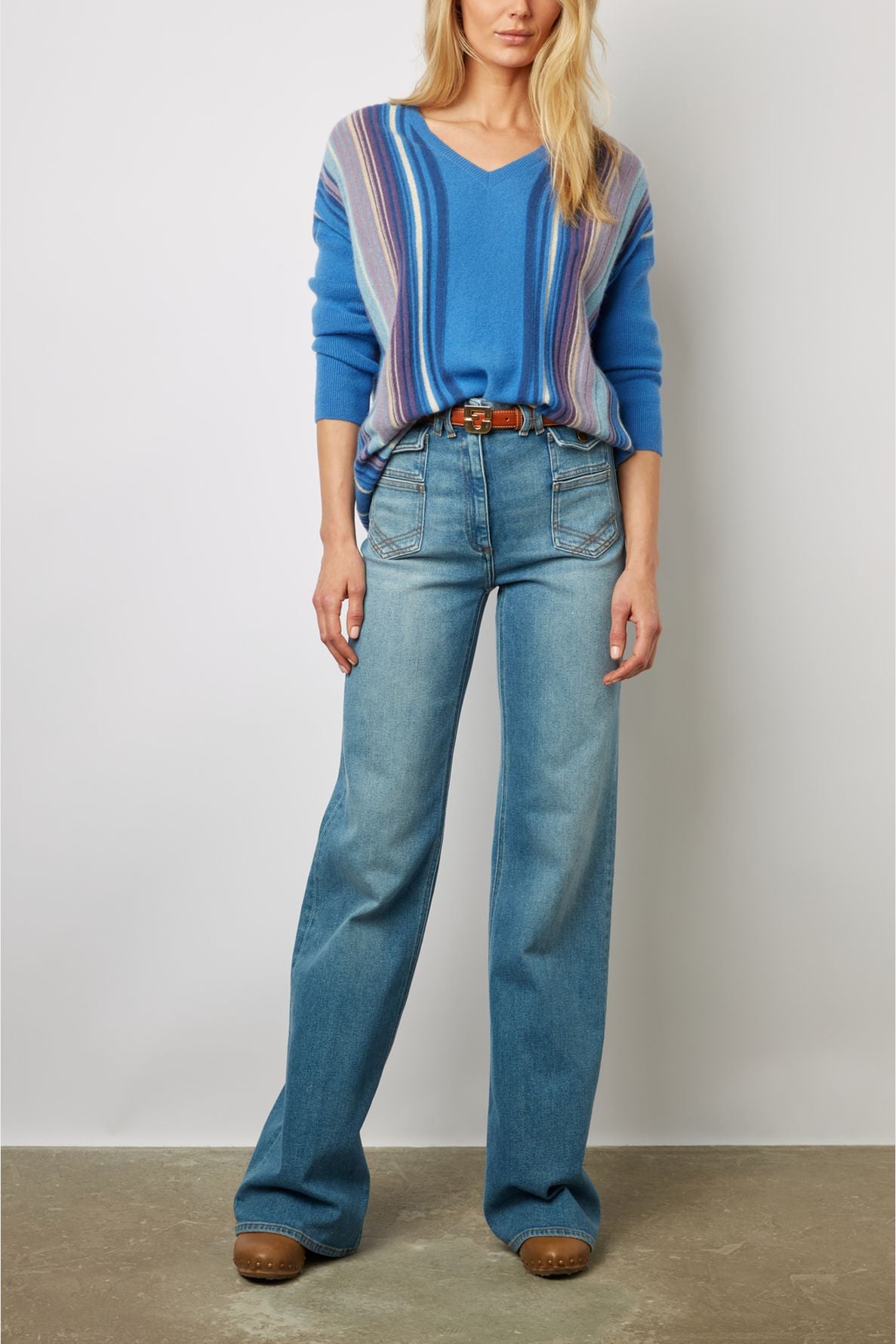 Woman wearing a blue rayé sweater tucked into high-waisted, wide-leg jeans with a brown belt and brown shoes, standing against a plain background for a subtle rétro look.