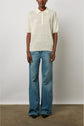 A person wearing a light short-sleeve polo shirt and wide-leg blue jeans stands against a plain white wall on a concrete floor.