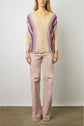A person stands against a plain background wearing a beige V-neck sweater with colorful vertical stripes and light pink pants.
