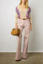 A woman stands against a plain wall, wearing a beige V-neck sweater with colorful stripes, light pink high-waisted trousers, and brown shoes. She holds a tan handbag in her left hand.