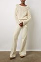 A person wearing a cream knit sweater and high-waisted, wide-leg cream pants stands against a plain wall, with hands clasped at the waist and brown heeled shoes visible.