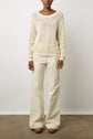 A person stands against a plain wall wearing a cream knit sweater, wide-leg cream pants, and brown shoes. Their hands rest at their sides, and the floor is bare concrete.