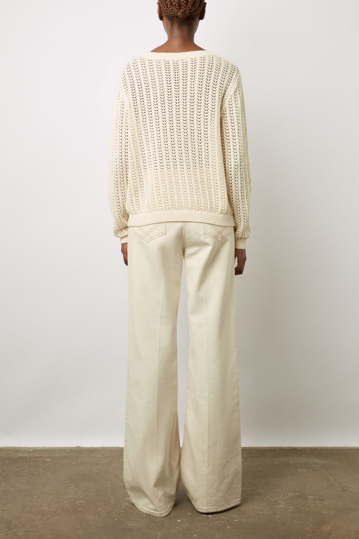 A person stands facing away, wearing a loose-knit cream sweater and high-waisted, wide-leg cream pants. The outfit and background are neutral, and the floor appears to be concrete.