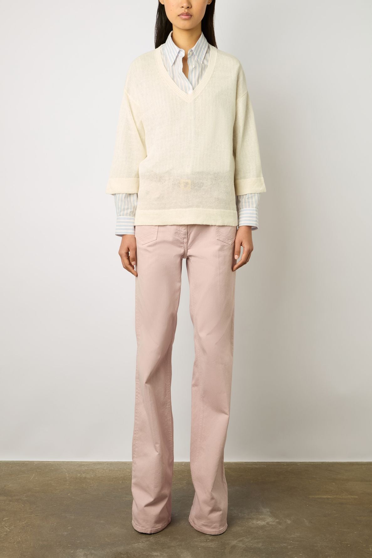 A person stands against a light wall wearing a cream V-neck sweater over a collared shirt with light pink wide-leg pants. Their hands are at their sides, and their head is partially out of frame.