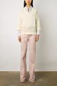 A person stands against a light wall wearing a cream V-neck sweater over a collared shirt with light pink wide-leg pants. Their hands are at their sides, and their head is partially out of frame.