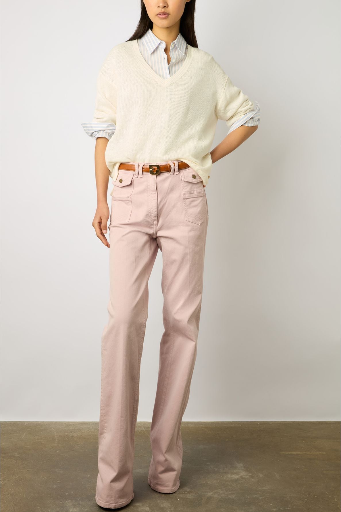 A person wearing a cream sweater over a blue striped shirt, pink high-waisted trousers, and a brown belt stands against a plain background. Their hand rests on their hip. Their face is not visible.