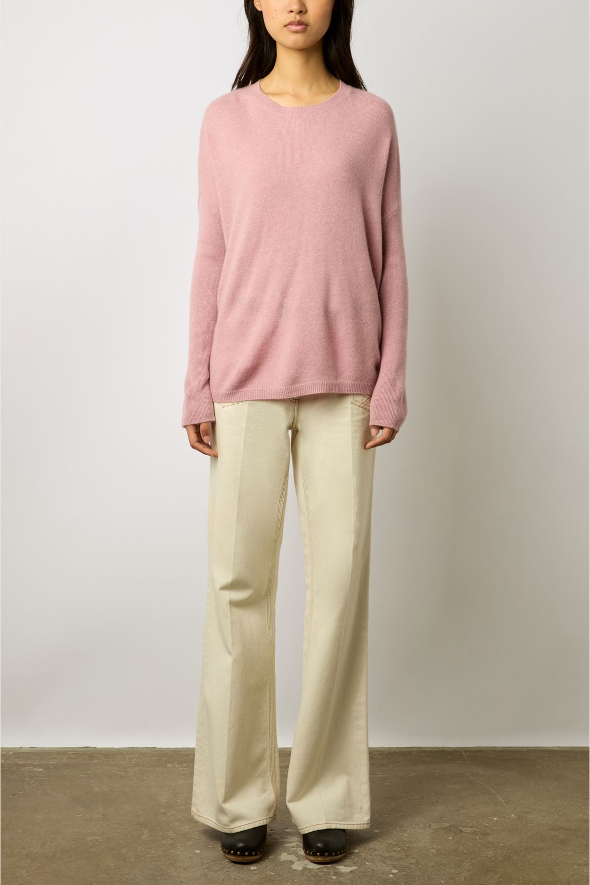 A woman wearing a loose pink sweater and wide-leg beige pants stands against a plain white wall, facing forward.