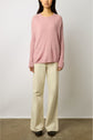 A woman wearing a loose pink sweater and wide-leg beige pants stands against a plain white wall, facing forward.