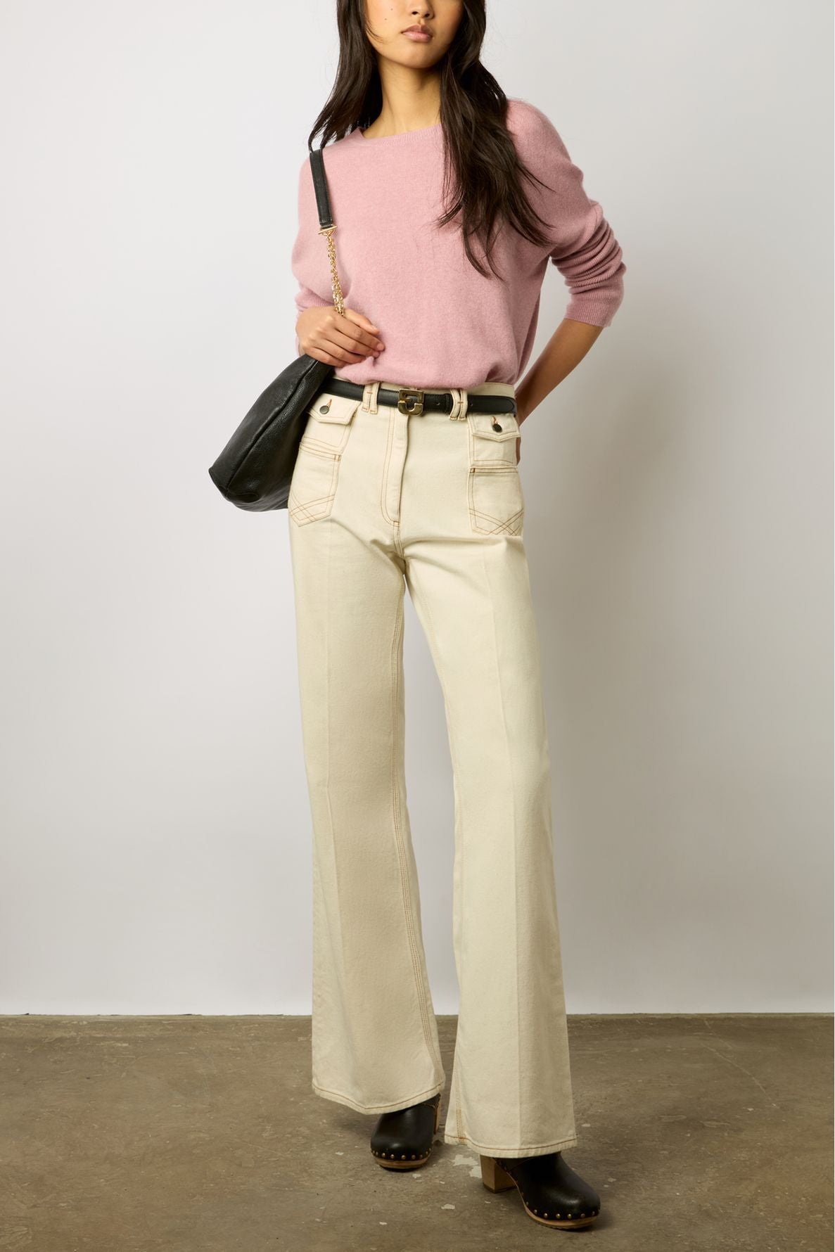 A woman stands against a plain wall, wearing a pink sweater, high-waisted cream wide-leg pants, black platform clogs, and a black shoulder bag. Her left hand is in her pocket. Her face is partially out of frame.