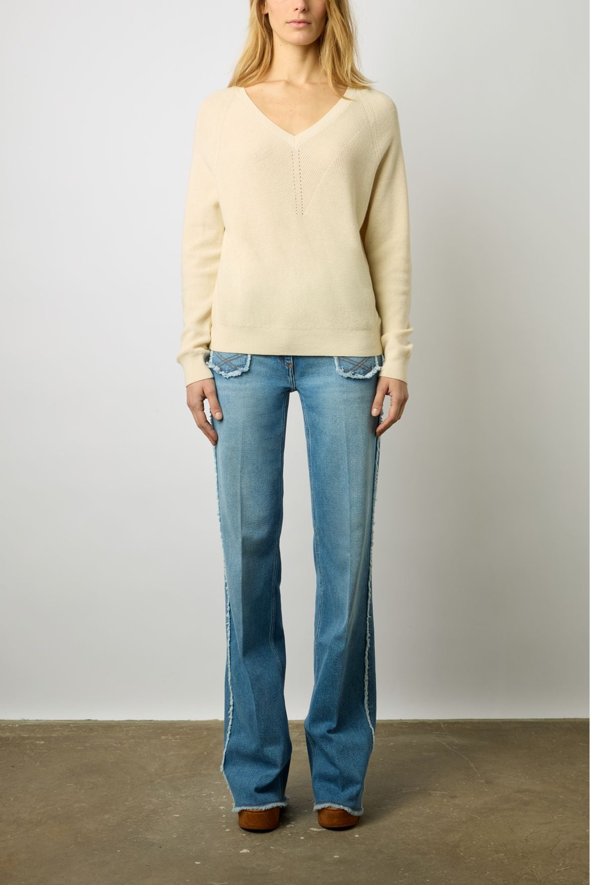 A woman stands against a plain wall wearing a cream V-neck sweater, light blue flared jeans with frayed seams, and brown shoes. Her hands rest by her sides, and her face is partially out of frame.