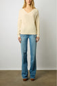 A woman stands against a plain wall wearing a cream V-neck sweater, light blue flared jeans with frayed seams, and brown shoes. Her hands rest by her sides, and her face is partially out of frame.