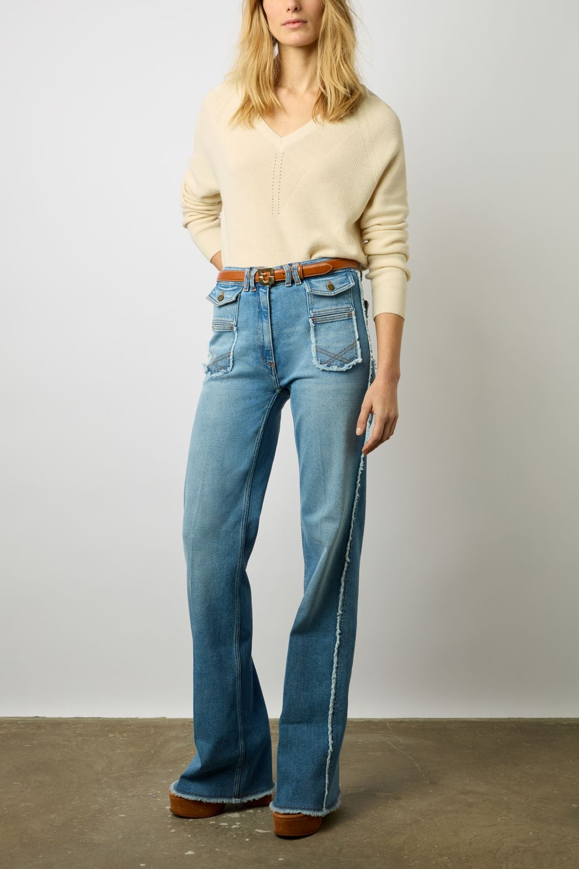 A person wearing a cream sweater, high-waisted wide-leg jeans with large front pockets, a brown belt, and brown platform shoes stands against a plain background.
