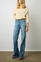 A person wearing a cream sweater, high-waisted wide-leg jeans with large front pockets, a brown belt, and brown platform shoes stands against a plain background.