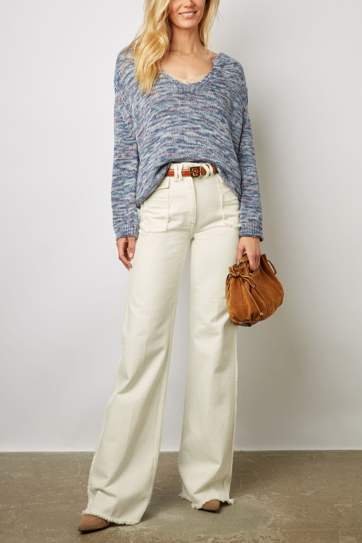 A woman wearing a blue pull chiné sweater, white wide-leg pants, tan belt, and tan pointed shoes holds a small brown suede bag. She stands against a plain background, smiling with loose, wavy blonde hair.