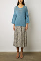 Woman wearing a loose blue knit sweater over a floral midi skirt and black strappy sandals, standing against a plain light background.