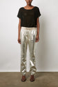 A person wearing a sheer black short-sleeve top, shiny metallic silver pants reminiscent of maille irisée, and black sandals stands against a plain white wall on a concrete floor.