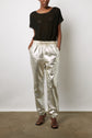 A person wearing a sheer black top, metallic silver pants, and black open-toe heels stands against a plain white wall with hands in pockets. The look is elevated by a pull léger femme en maille irisée, adding subtle shimmer and effortless elegance to the ensemble.
