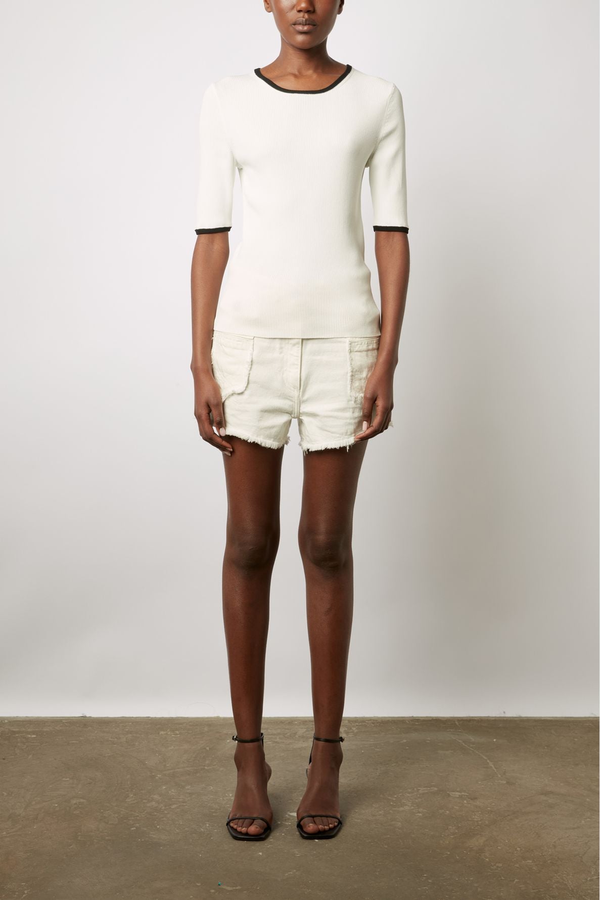 A woman stands against a plain background wearing a white short-sleeved top with black trim, white shorts, and black high-heeled sandals. Her arms are relaxed by her sides.