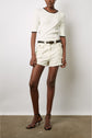 A person wearing a white short-sleeve top, cream-colored belted shorts, and black heeled sandals stands against a plain white wall and concrete floor.