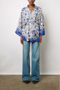 Person wearing a blue floral wrap blouse reminiscent of a kimono en soie with a belt, paired with wide-leg blue jeans and black studded shoes, standing against a plain white background.