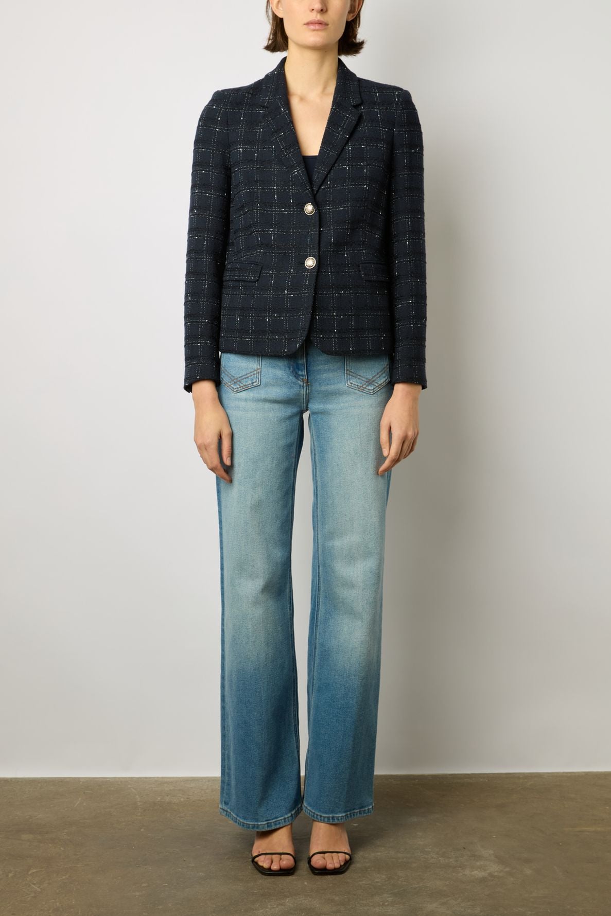 Woman wearing a dark plaid blazer, light blue wide-leg jeans, and black open-toe heels standing against a plain light-colored wall.